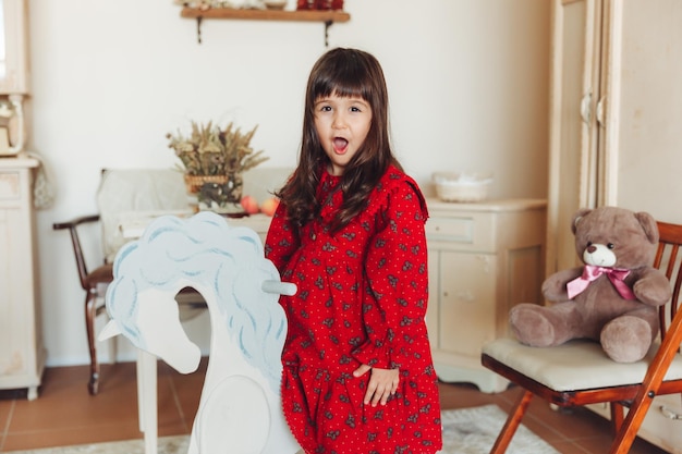 A charming little girl in a dress made of natural material rides a rocking horse in her room antique nursery interior