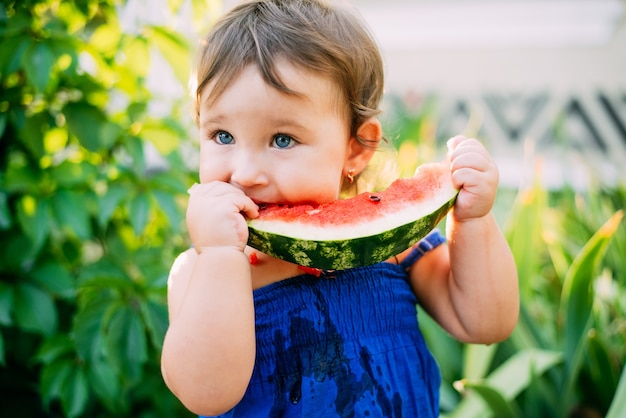 Charming little girl eating watermelon in the yard on the background of plants, very cute