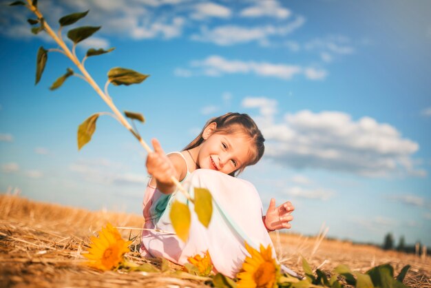 Charming little girl holding a sunflower in hand sitting on the field on a hot sunny summer day