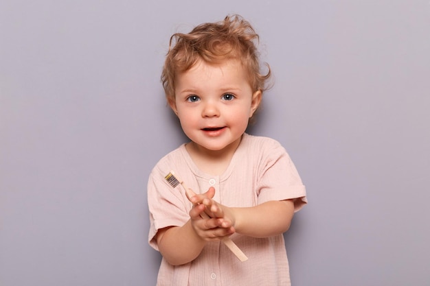 Charming little girl holding toothbrush isolated on gray background looking at camera holding toothbrush trying to brush teeth by herself