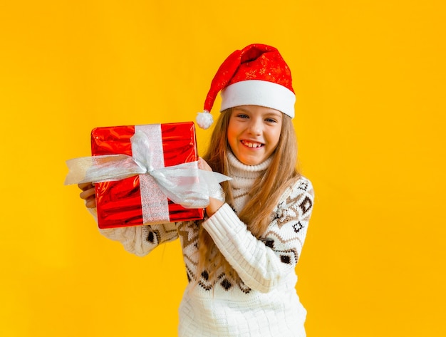 Charming little girl in a knitted sweater and Santa hat with a gift on a yellow background.