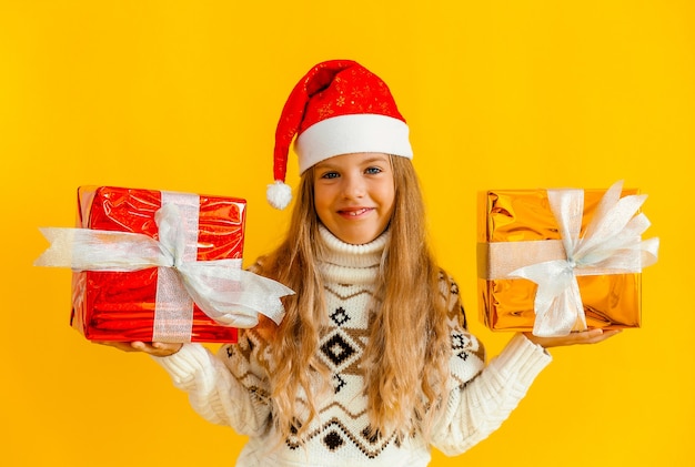 Charming little girl in a knitted sweater and Santa hat with a gift on a yellow background.