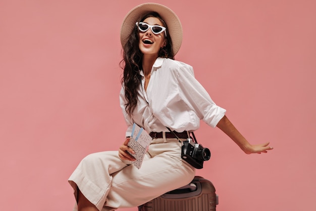 Charming long curly haired lady in cool sunglasses, modern clothes and light hat posing with camera and tickets on pink wall