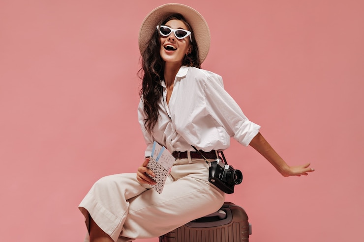 Photo charming long curly haired lady in cool sunglasses, modern clothes and light hat posing with camera and tickets on pink wall