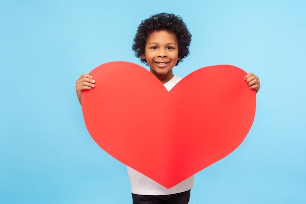 Charming lovely cute little boy with curly hair looking romantic and handsome with big red paper heart, child holding symbol of love, charity, affection. indoor studio shot isolated on blue background