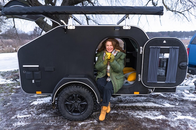 Charming mature woman enjoying her coffee in camper trailer dressed up warmly camper parked at the frozen river to enjoy time early spring time Vacation and journey concept