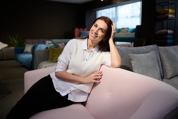 Charming middle aged European woman interior designer business owner sitting on sofa in furniture store and smiling beautiful smile looking at camera