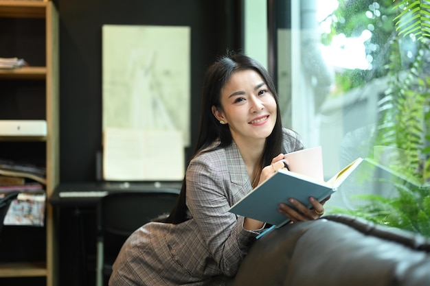 Charming millennial businesswoman resting taking a break from work and reading a book on sofa