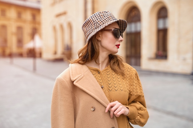 가을 베이지 색 코트와 선글라스에 매력적인 모델 소녀, 얼굴로 서있는 격자 무늬 파나마 모자