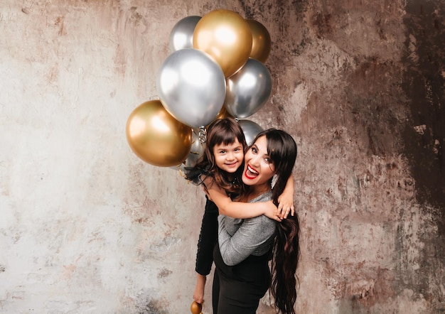 Charming mom and little daughter in the same outfit pose together after a birthday party Portrait of a charming girl hugging her daughter balloons