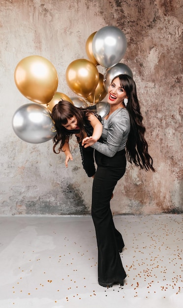 Charming mom and little daughter in the same outfit pose together after a birthday party Portrait of a charming girl hugging her daughter balloons