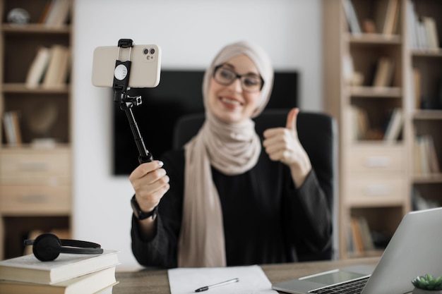 Очаровательная мусульманка в хиджабе разговаривает и жестикулирует во время записи видео на современный телефон на палочке