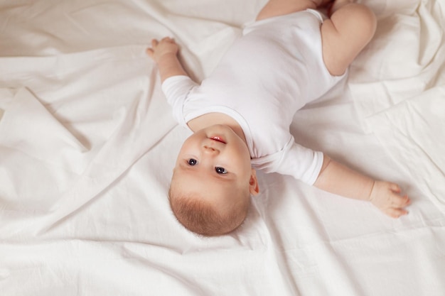 흰색 바디수트를 입은 매력적인 신생아는 흰색 천으로 된 위쪽 전망에 등을 대고 누워 있습니다.