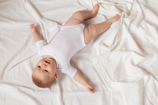Очаровательный новорожденный ребенок в белом боди лежит на спине на белой ткани, вид сверху