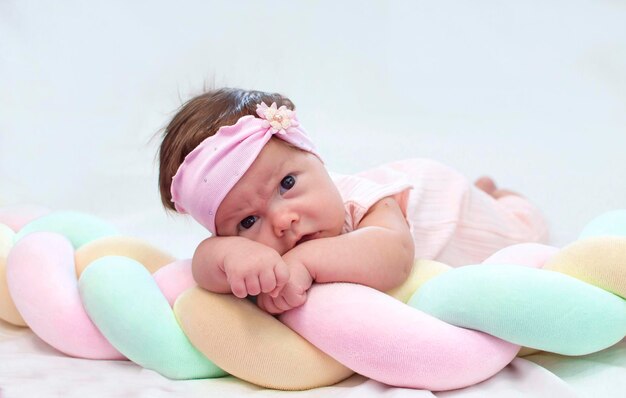 Charming newborn baby with bow on her head is lying on her stomach on a white bed at home looking into the camera morning of the child Concept of children's goods Happy childhood of healthy child