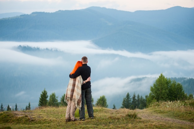 Фото Очаровательная пара стоит на холме, наслаждаясь утренней дымкой над горами. молодая пара, обнимая друг друга. рыжеволосая девушка покрыта одеялом. вид сзади