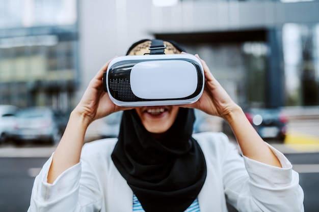 Charming positive smiling stylish muslim woman standing outdoors and putting vr headset on. Millennial generation.