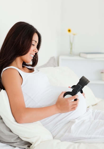 Charming pregnant woman putting headphones on her belly while lying on a bed