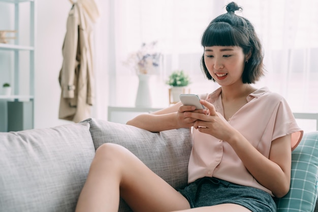 Charming pretty asian korean girl sitting on sofa in modern living room at home doing online shopping through internet. beautiful lady having smart phone in hands relaxing on couch in bright morning