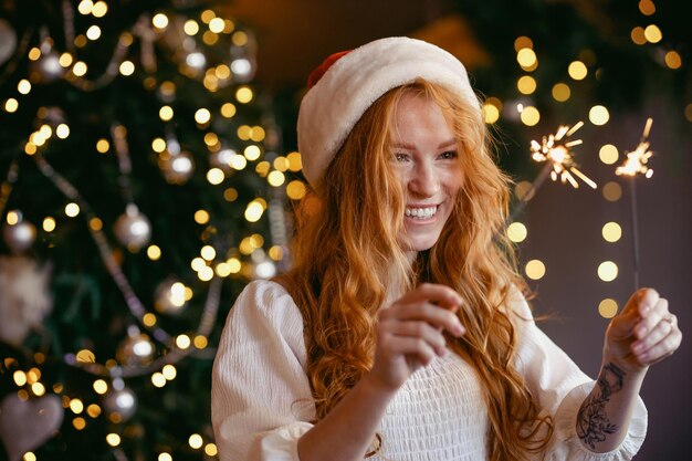 Charming red-haired girl in a santa hat with sparklers in her hands smiling at the camera.