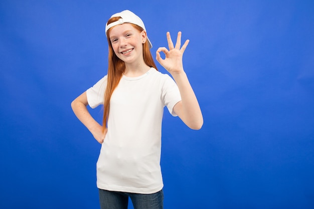 Charming red-haired teenager girl in a white T-shirt shows a blank print space on a blue studio background