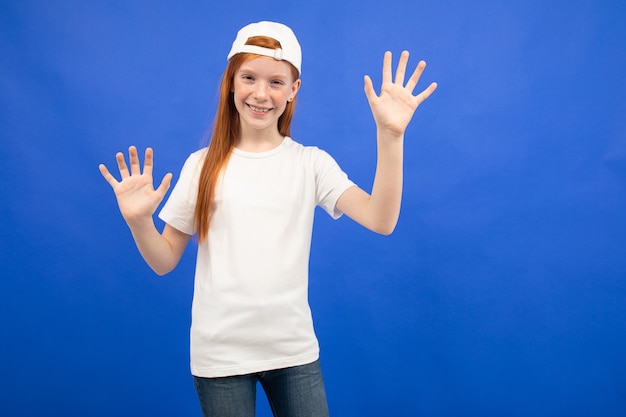 Charming red-haired teenager girl in a white T-shirt with a layout shows ten fingers on a blue studio background.