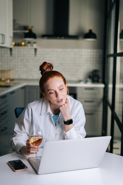 Charming redhead young woman having video call conversation using laptop and drinking champagne. Happy lady talking via webcam gathering friends, boyfriend sending online congratulations.