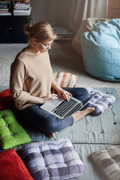 Affascinante ragazza caucasica rilassata con gli occhiali con i capelli biondi, seduta sul pavimento con i piedi incrociati, tenendo il laptop mentre si lavora con il notebook o la messaggistica, guardando video in internet.