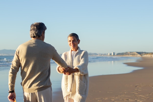 Очаровательная пожилая пара развлекается на берегу моря. Счастливая короткошерстная дама держит своего мужа за руку, наслаждаясь теплым осенним вечером на пляже вместе. Выход на пенсию, концепция досуга
