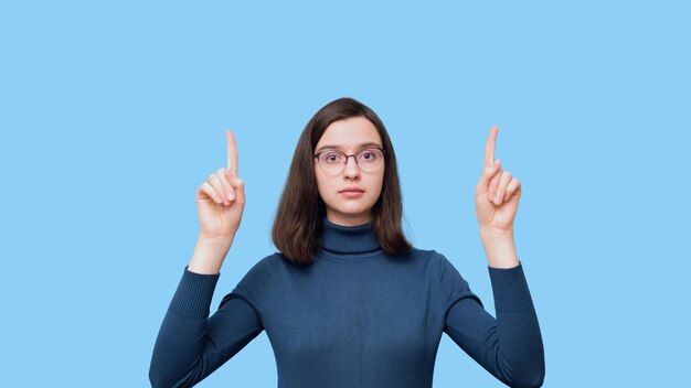 A charming serious student girl with glasses in a blue sweater raised her hands up pointing to an empty space