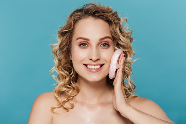 charming shirtless woman 20s with curly blond hair applying makeup with powder puff isolated over blue wall