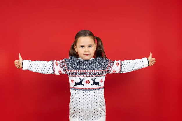 Affascinante ragazza di sei anni in maglione natalizio bianco con renne che alzano i pollici e con le braccia alzate...