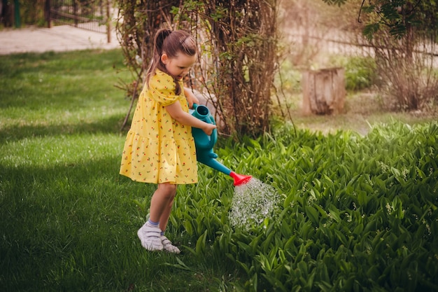 Charming sixyearold girl in a yellow dress watering lilies of the valley from a blue watering can a ...