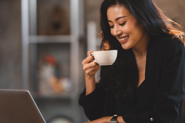 매력적인 똑똑한 아시아 비즈니스 여성 기업가는 노트북을 사용하여 교육 회사 경력 개발 세미나 앉아서 로프트 사무실이나 카페 프리랜서 직업 생활 방식에서 테이크아웃 머그 뜨거운 음료를 들고 일합니다.
