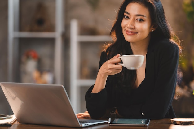 L'affascinante imprenditrice asiatica d'affari usa il laptop per lavorare la società di formazione professionale seminario di sviluppo della carriera sedersi al tavolo tenere la tazza da asporto bevanda calda in ufficio loft o caffè freelance stile di vita di lavoro