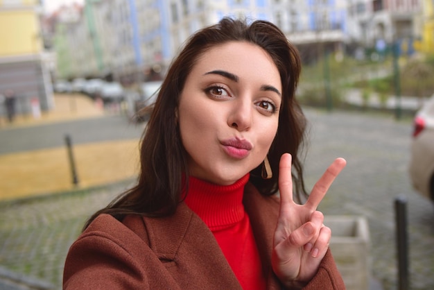 Photo charming smiling lady in a brown autumn coat takes a selfie on the street with a peace sign