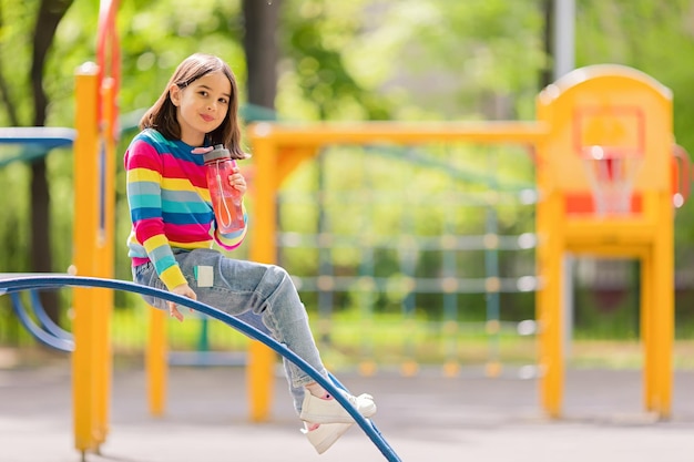 Фото Очаровательная улыбающаяся маленькая девочка сидит с пластиковой бутылкой воды на детской площадке