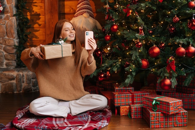 Charming smiling woman showing festive interior christmas tree and xmas gift box during video call