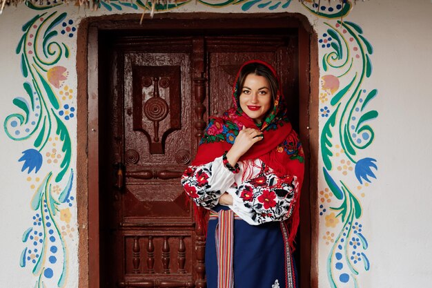 Charming smiling woman in traditional ukrainian handkerchief necklace and embroidered dress standing at background of decorated hut Ukraine style folk ethnic culture