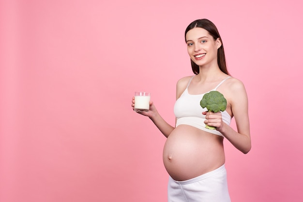 A charming smiling young pregnant woman, brown-haired, in a white top and white leggings, holds a glass of milk and broccoli in her hand. Diet concept for pregnant women, toxicosis.