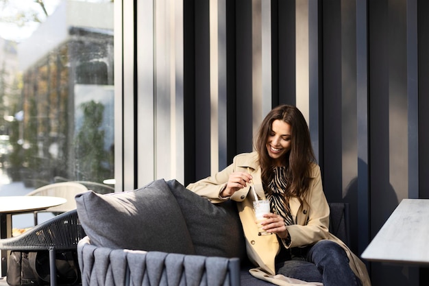 Charming smiling young woman sitting on a sofa drinking a drink on a terrace in a cafe Attractive girl resting outdoors