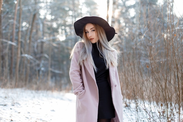 Charming stylish young woman in a luxurious hat in a pink coat in a knitted golf in a skirt posing in the winter forest 