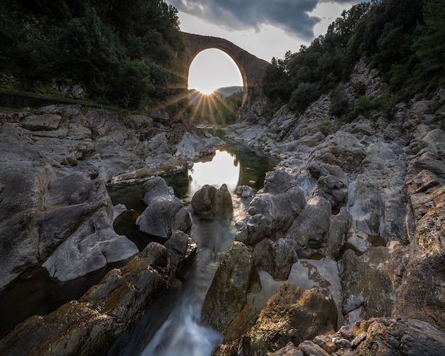 Charming sunset sun stars inside a bridge behind a small river surrounded by rocks with reflected warm light in Spain