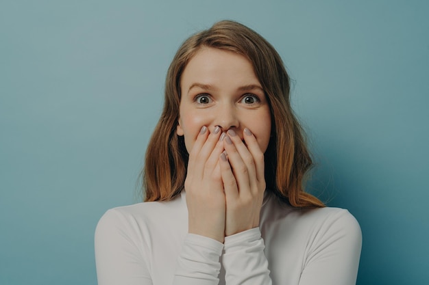 Photo charming surprised positive young woman closing her mouth with both hands in excitement