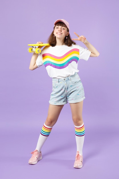 Charming teen girl in vivid clothes holding yellow skateboard, showing victory sign isolated on violet pastel wall background in studio. People sincere emotions, lifestyle concept. Mock up copy space.