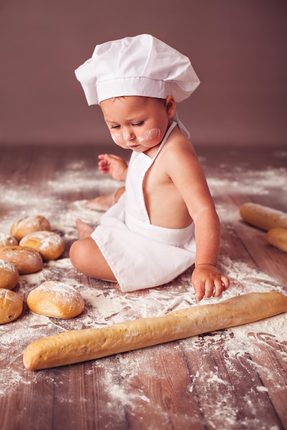 写真 バゲットで遊ぶパンの塊と小麦粉に座っている料理人とエプロンの帽子をかぶった魅力的な幼児の赤ちゃん