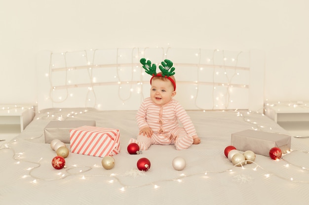 Charming toddler girl sitting on Christmas decorations bed and wearing striped long sleeve baby sleeper and festive deer horns, looking away with charming smile.