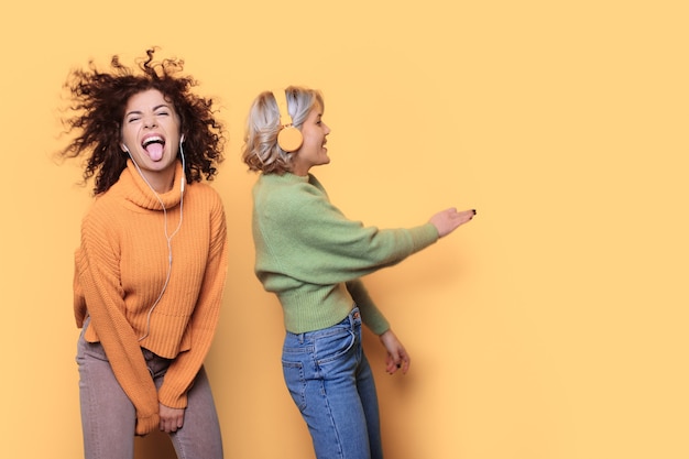 Photo charming two women dancing on a yellow wall with free space while listening to music