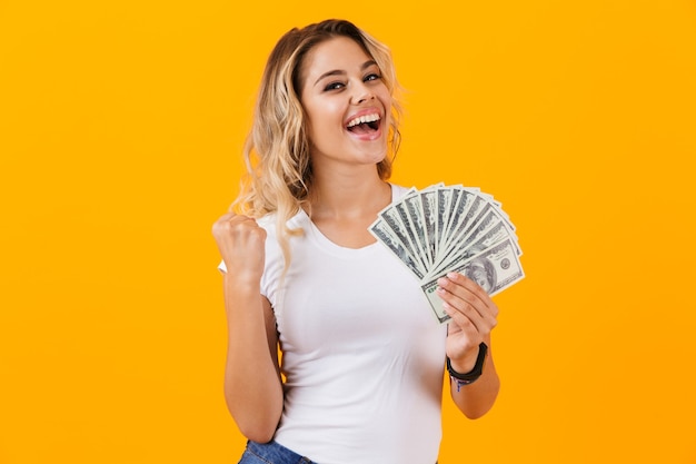 Charming woman in basic clothing holding fan of dollar money, isolated over yellow wall