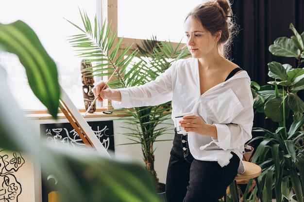 Affascinante donna in abiti casual seduta in uno studio d'arte e che usa il cavalletto per disegnare